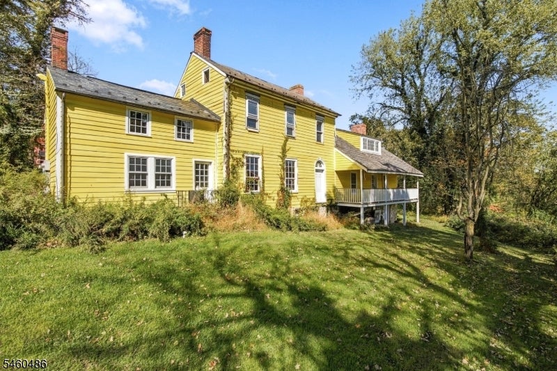 Yellow two-story house with brick chimneys and a raised covered deck, surrounded by a lawn and mature trees.