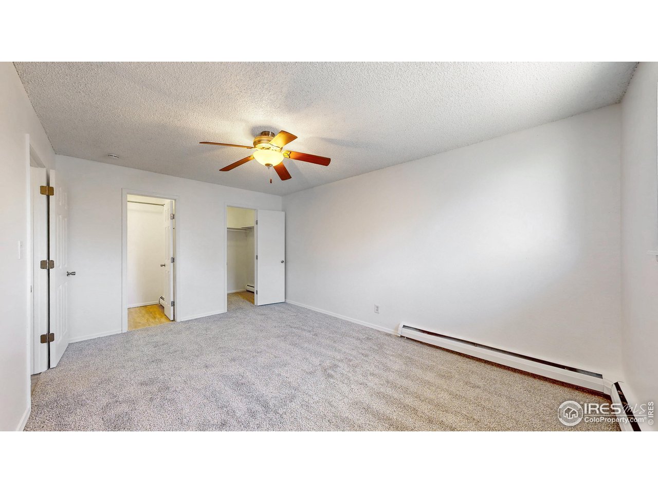 Empty carpeted bedroom with a lit ceiling fan, open closet, and long baseboard heater.