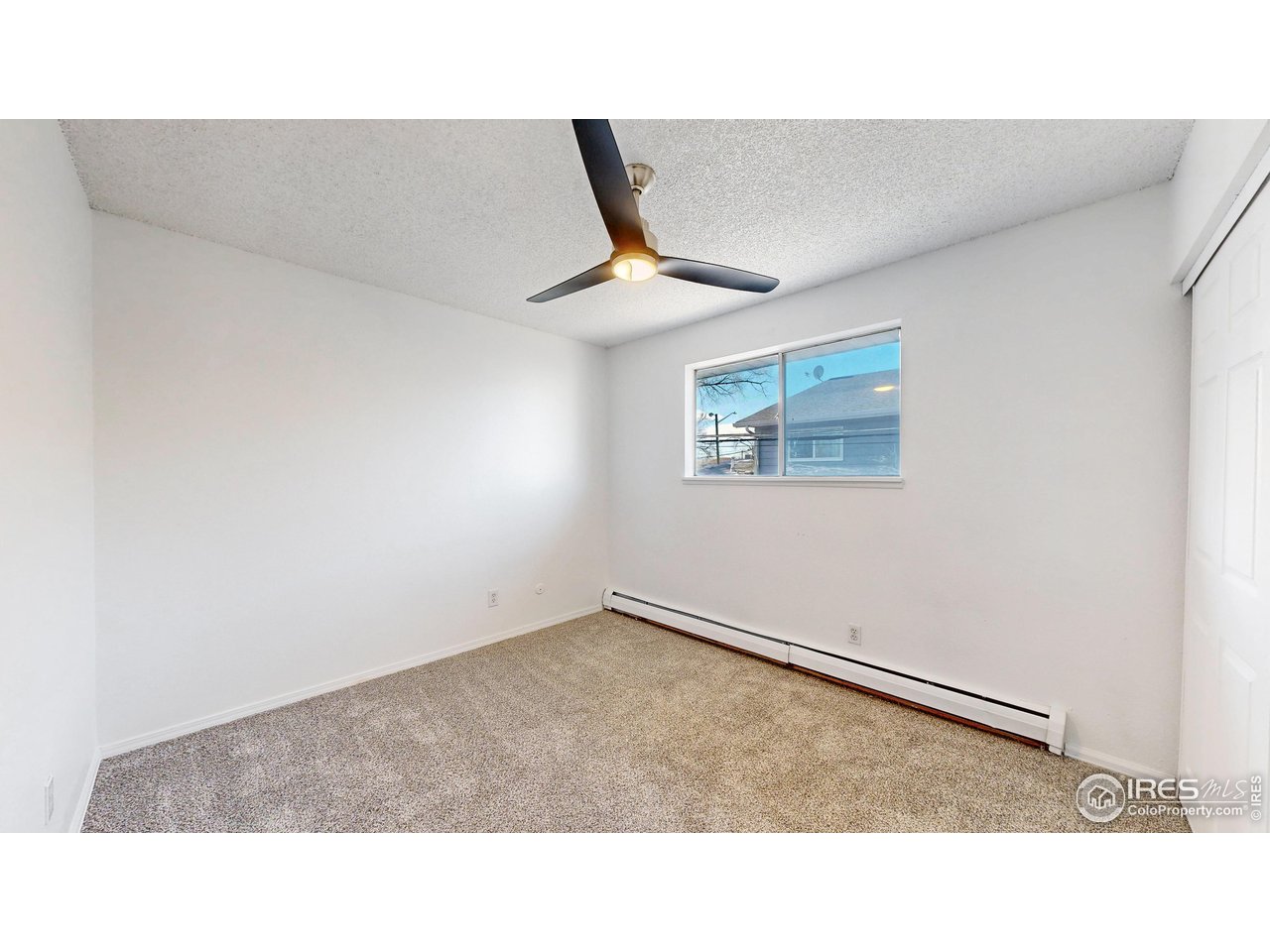 Empty bedroom with beige carpet, a black ceiling fan, and a window above a baseboard heater.