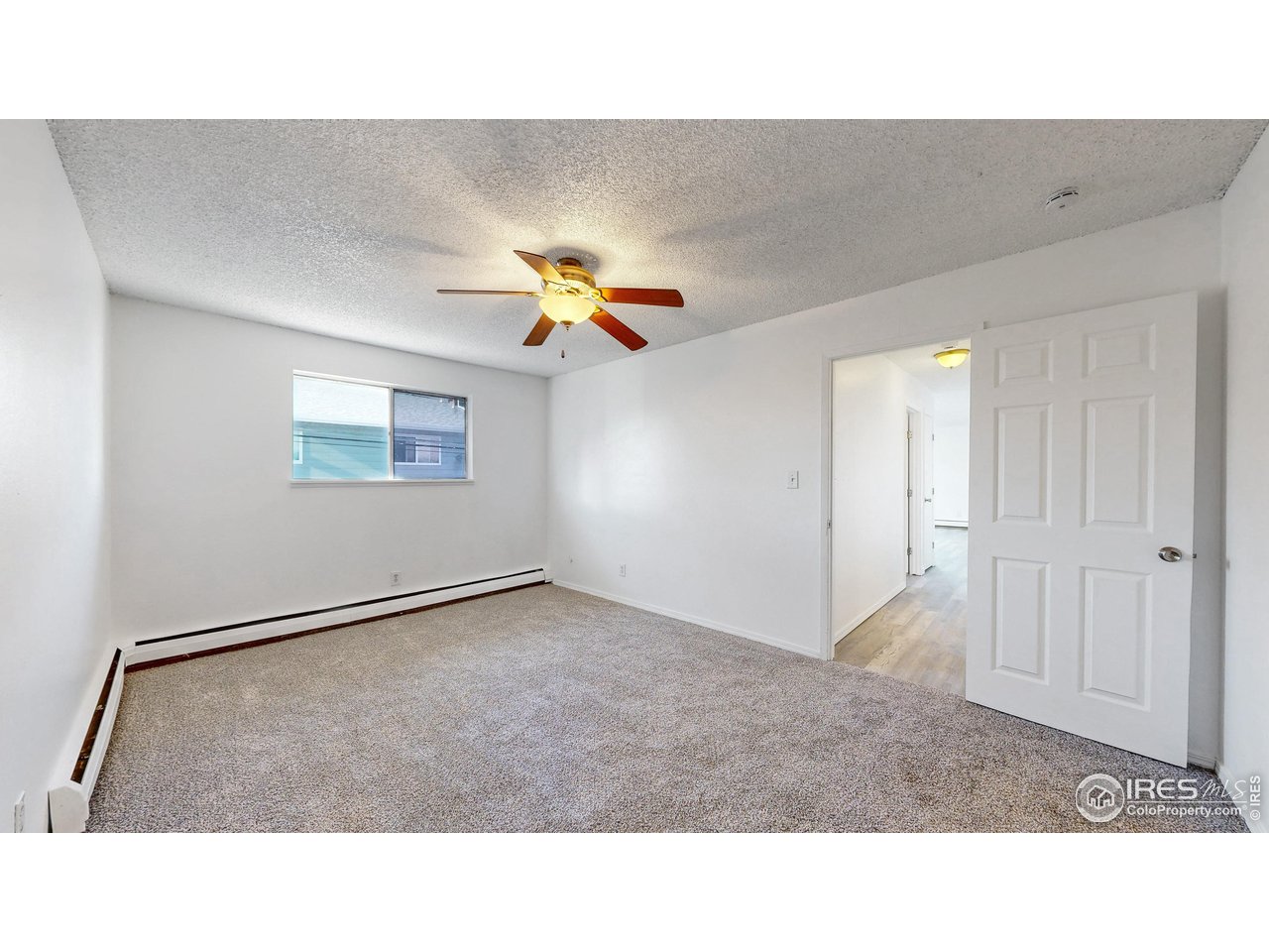 Empty carpeted bedroom with a ceiling fan, window, and open doorway to a wood-floored hall.