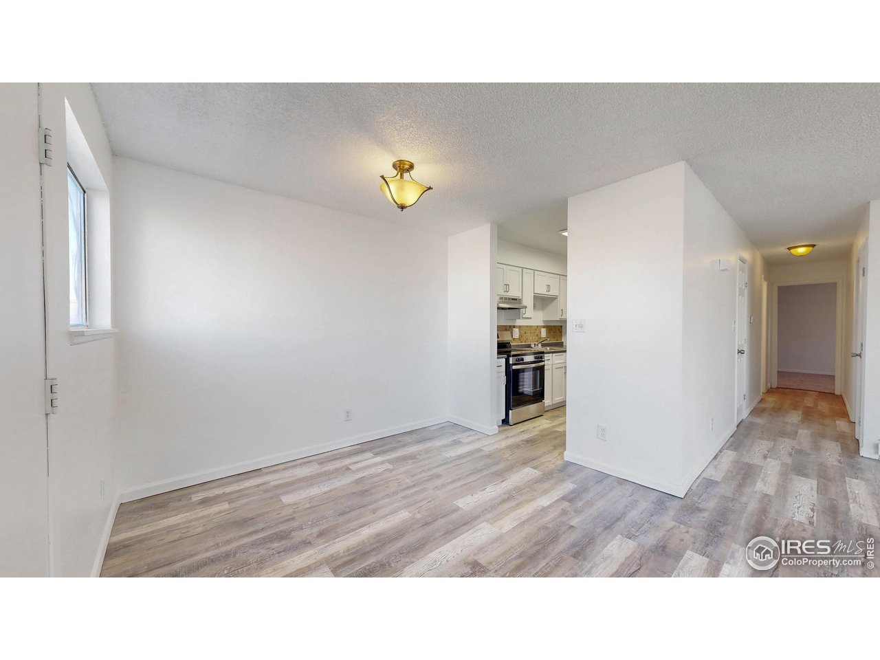 Empty living area with gray wood-look flooring, a window, and an open view to the kitchen and hallway.