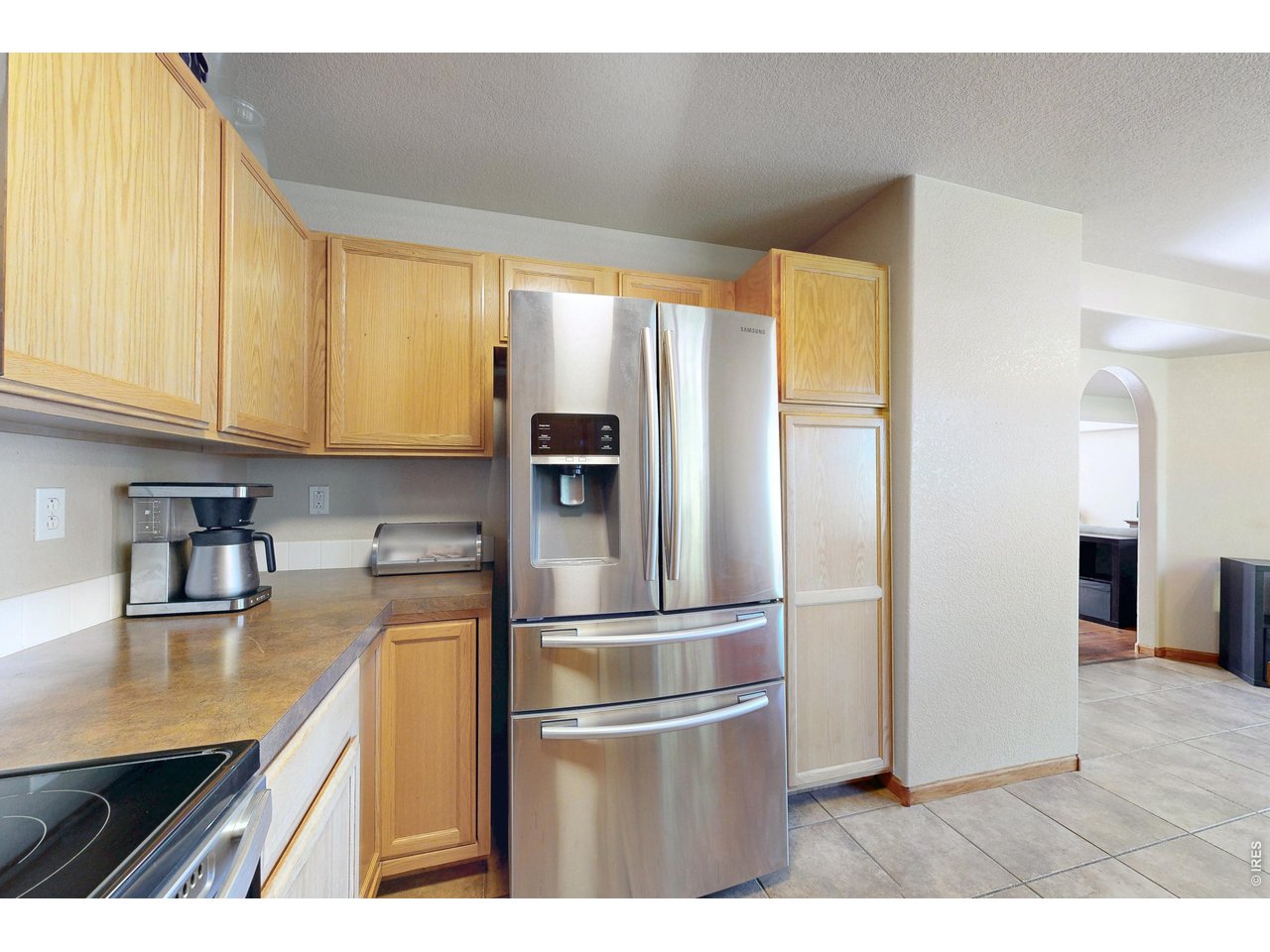 Kitchen with light wood cabinets, tan countertops, and a stainless steel refrigerator beside an arched opening.