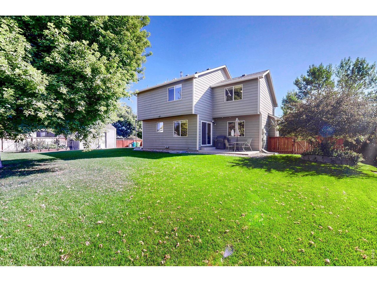 Two-story gray home behind a broad lawn, with mature trees and a fenced patio seating area.