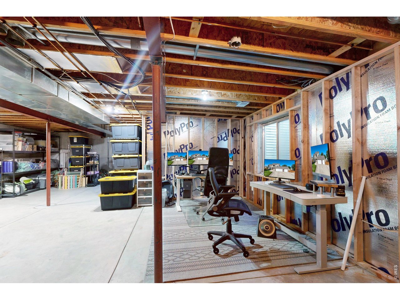 Basement office workspace with computer monitors, an office chair, exposed framing, and unfinished insulated walls.