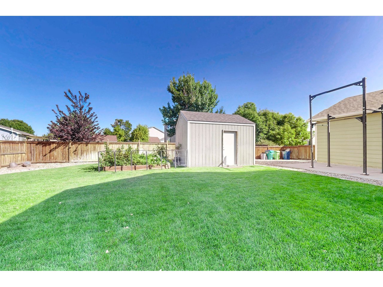 Fenced backyard with a large lawn, raised garden beds, gray storage shed, and paved area.