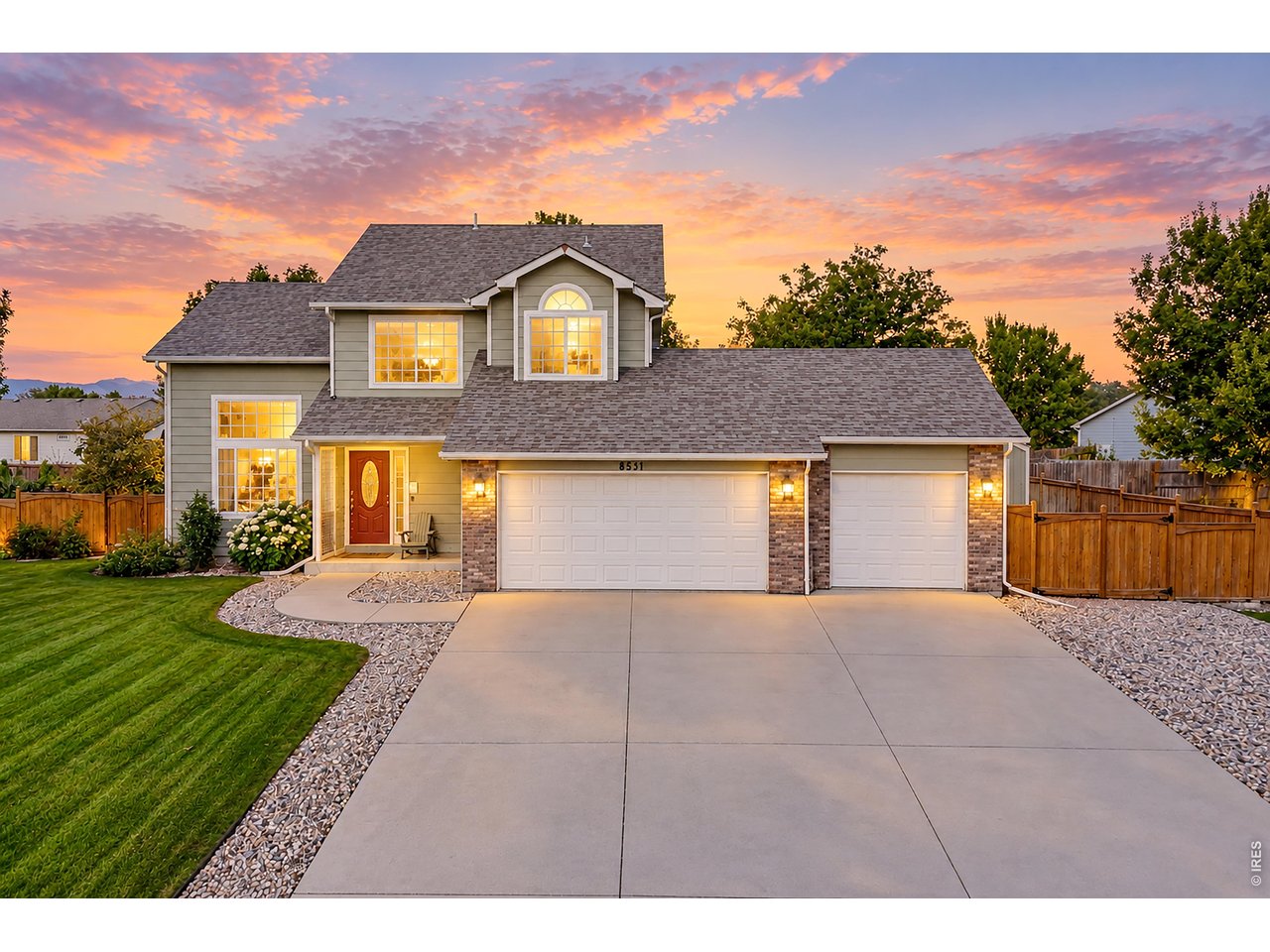 Two-story gray home with a three-car garage, red front door, and illuminated windows at sunset.