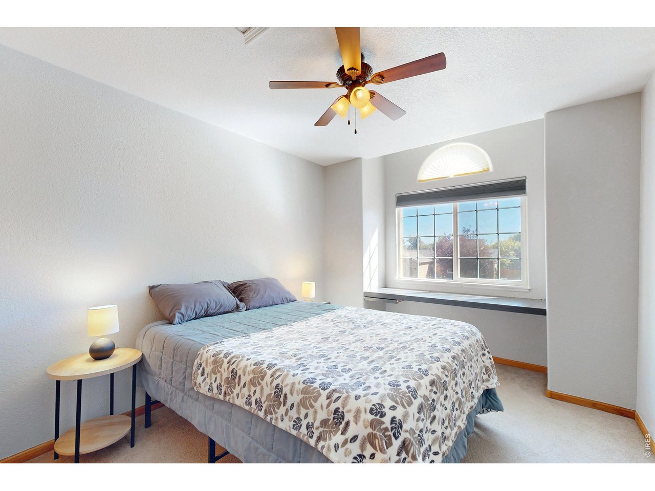 Furnished bedroom with a floral quilt, ceiling fan, and large window with a half-round transom.