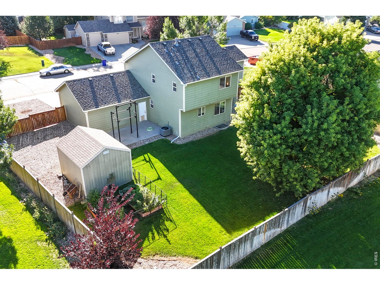 Aerial view of a fenced backyard with a wide lawn, storage shed, and pergola beside a two-story house.