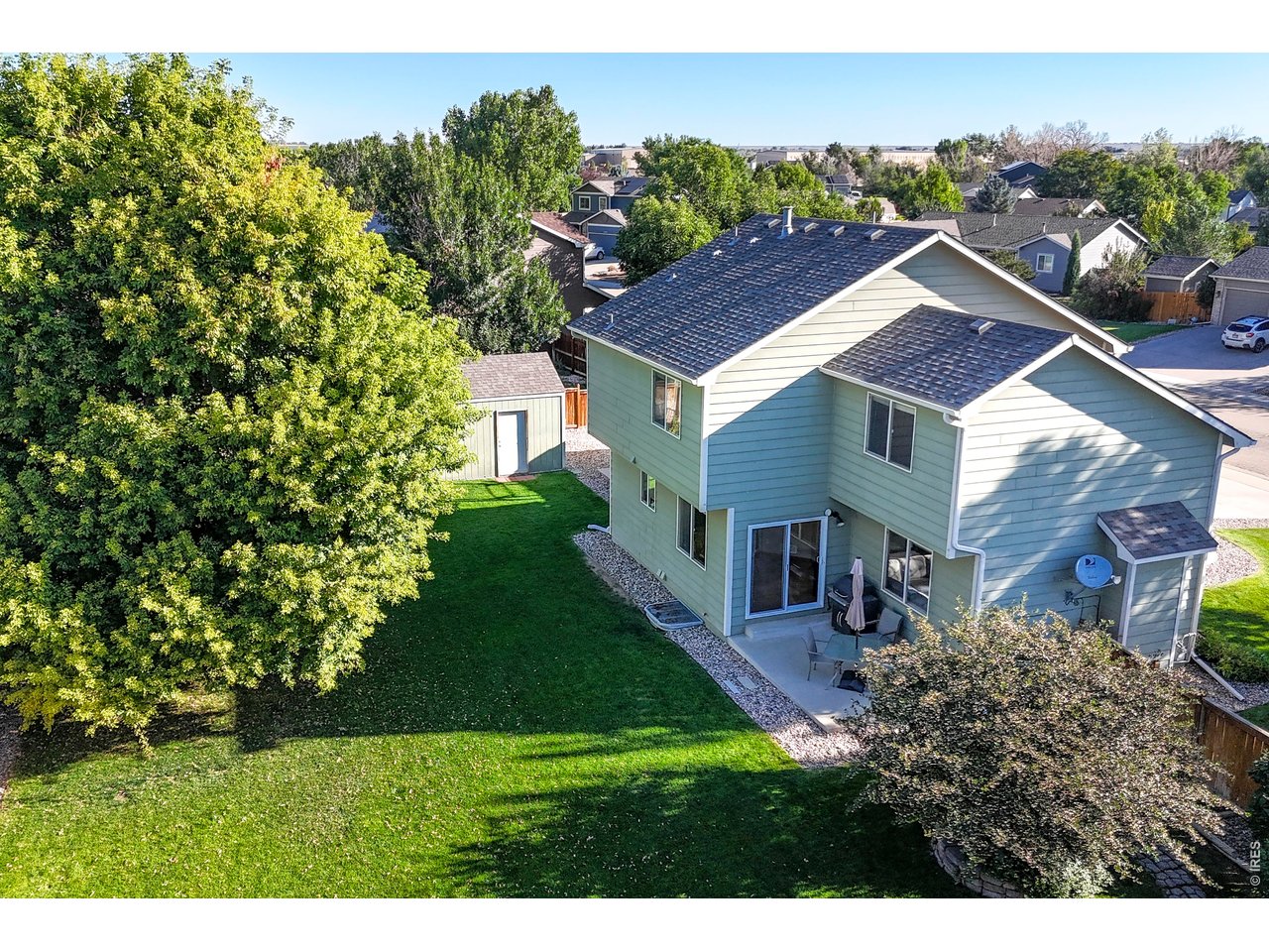 Aerial view of a light-green two-story home, patio, lawn, mature trees, and backyard shed.