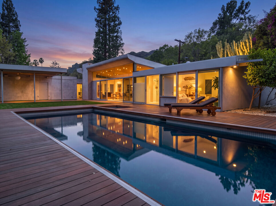 Modern glass-walled home with illuminated interiors and a reflecting pool at dusk.