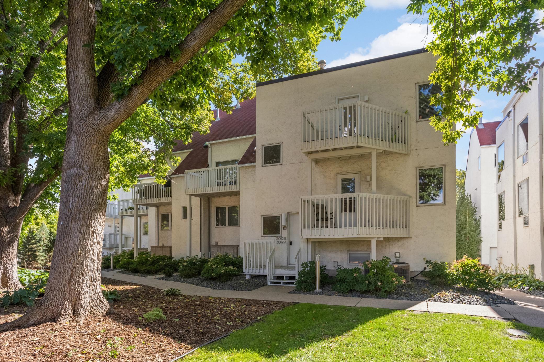 Beige multi-unit building with balconies, framed by a large leafy tree and landscaped grounds.