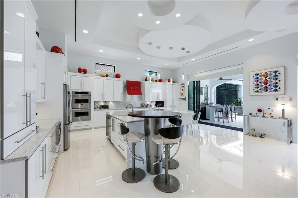 Modern white kitchen with a round island, bar stools, glossy cabinetry, and a sculptural ceiling.