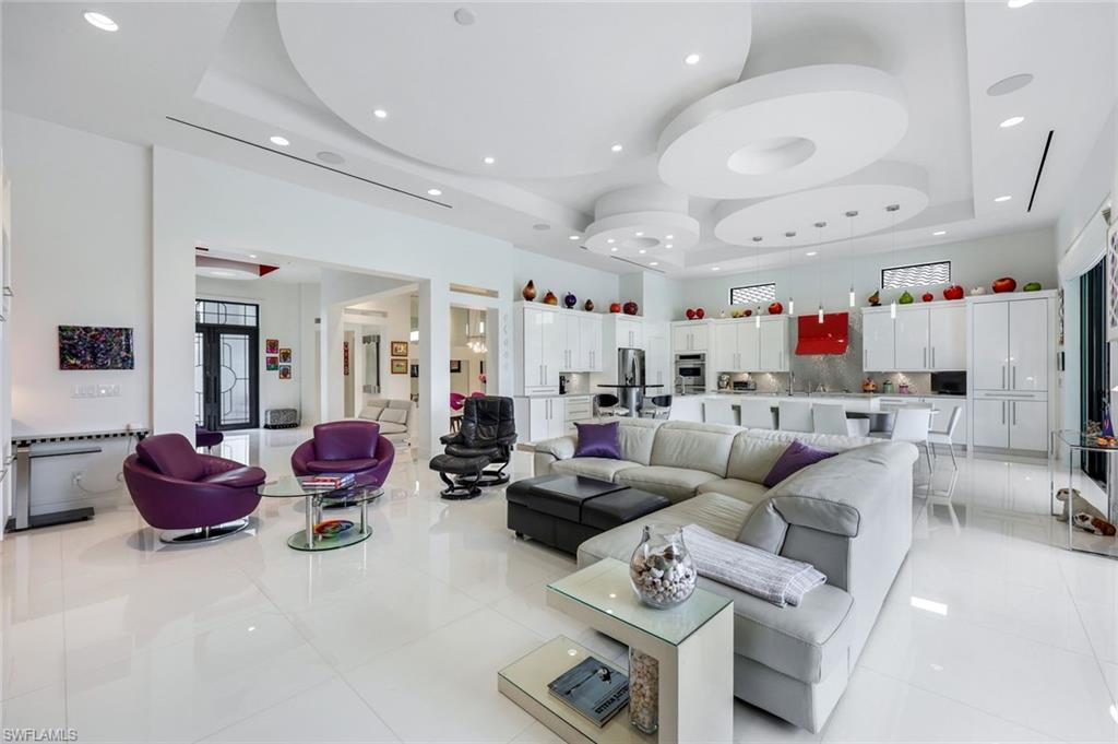 Open living room with a gray sectional, purple chairs, and a sculptural ceiling beside the white kitchen.