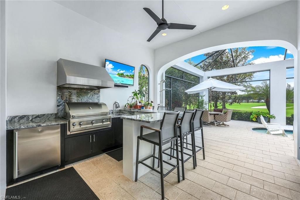 Covered outdoor kitchen with a stainless-steel grill and bar overlooking a furnished patio and golf course.