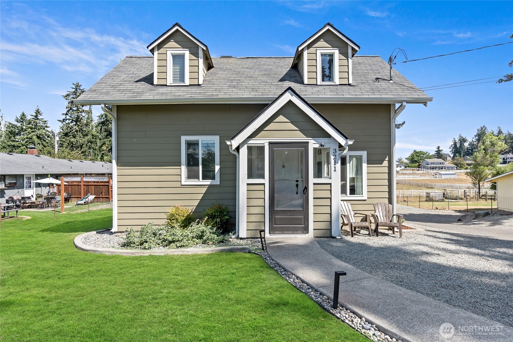 Gray-green two-story house with twin dormers, a covered entry, lawn, and gravel driveway.