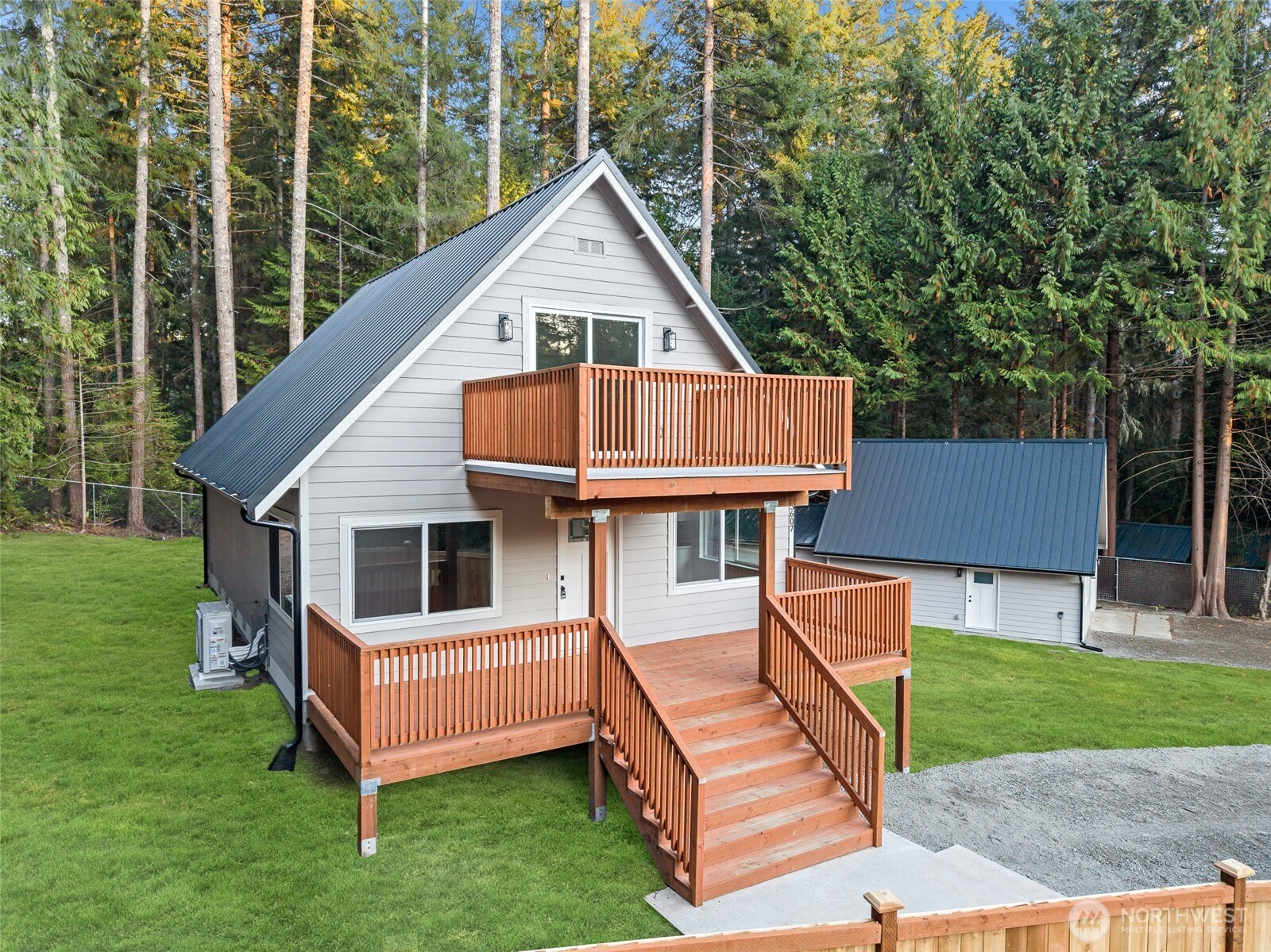 Gray A-frame house with two wooden decks and stairs, surrounded by evergreen trees.