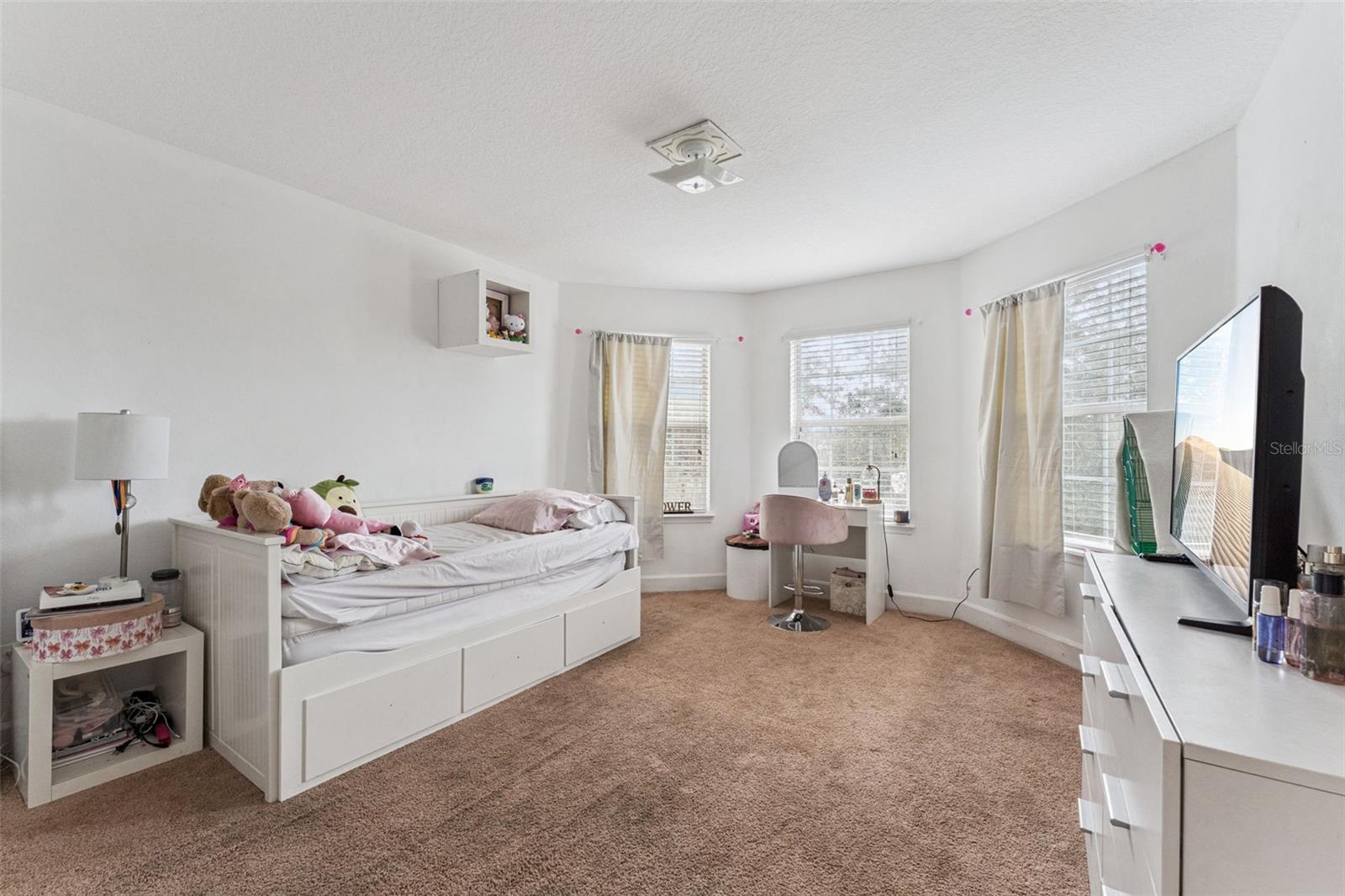 Bedroom with a white daybed, plush toys, beige carpet, and a vanity by three curtained windows.