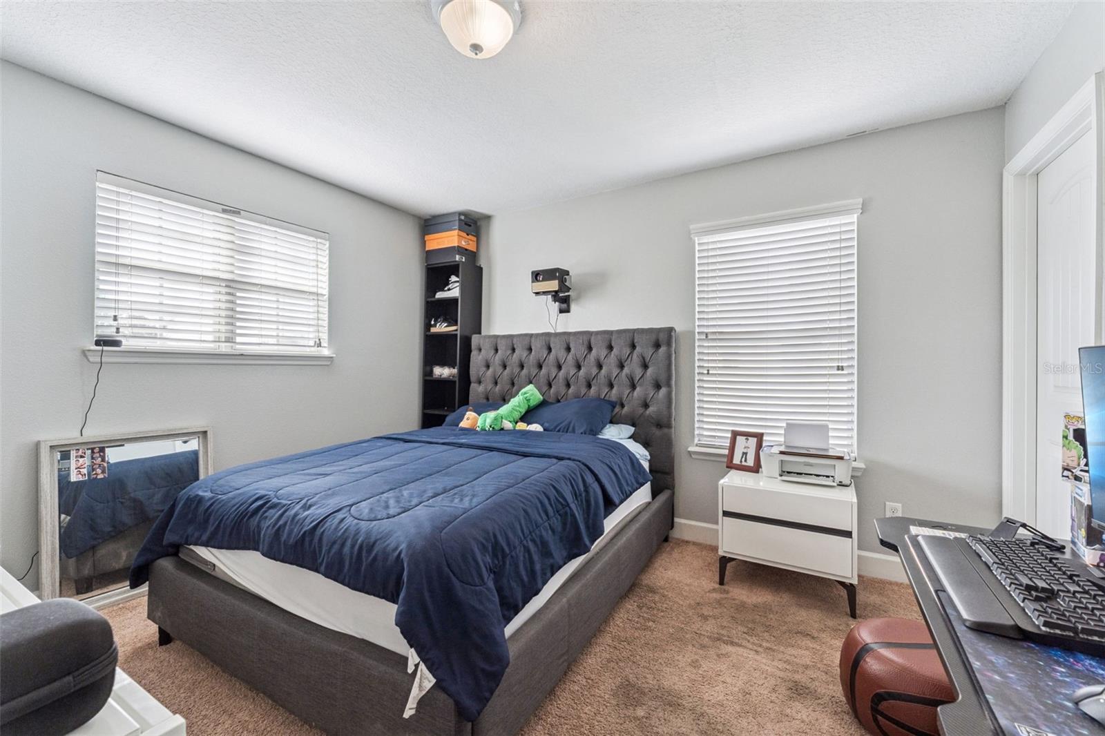 Bedroom with a gray tufted bed, navy bedding, and a desk beside two windows.