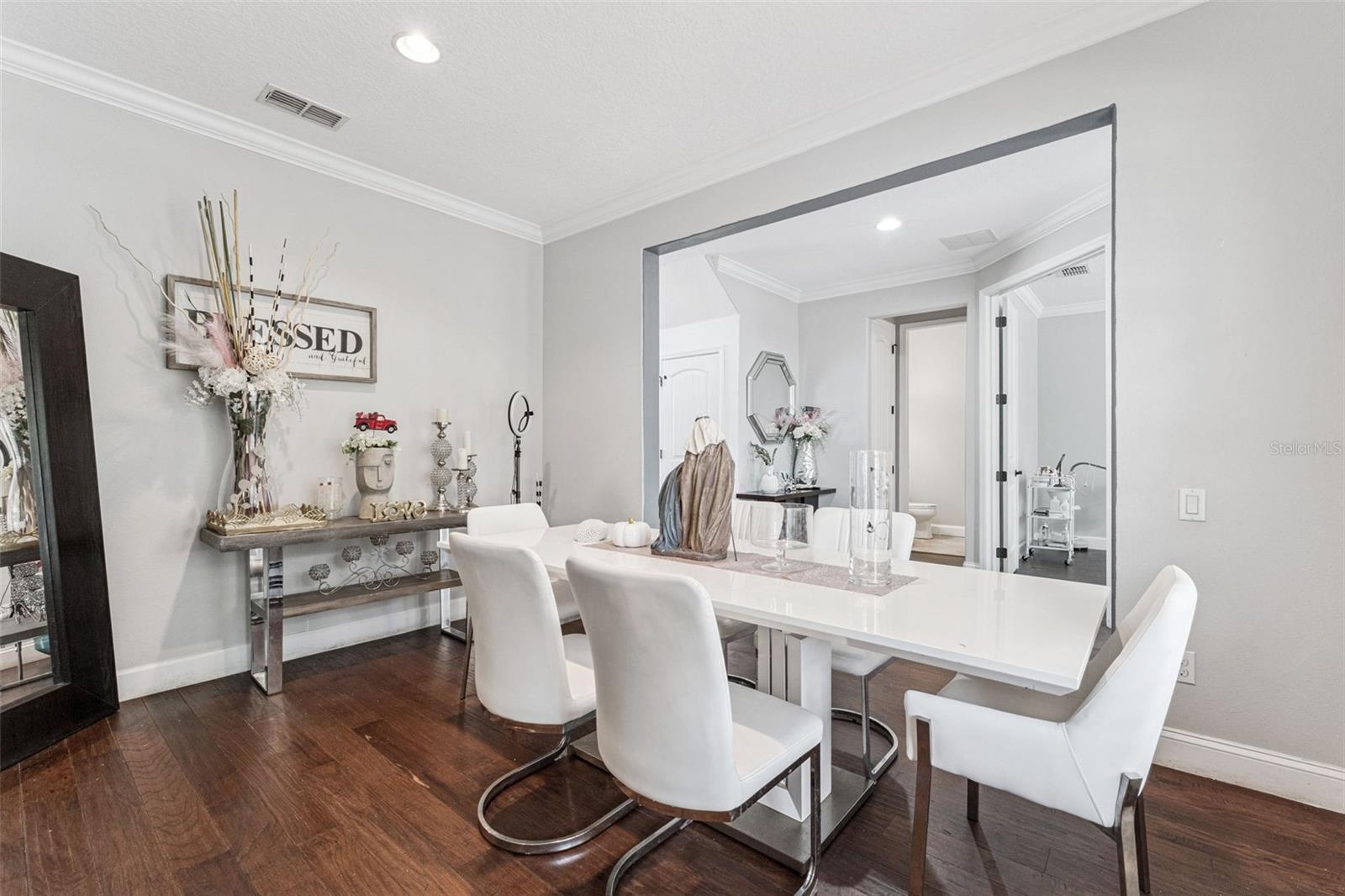 Staged dining room with a white table, upholstered chairs, dark wood floors, and a console beneath a “BLESSED” sign.