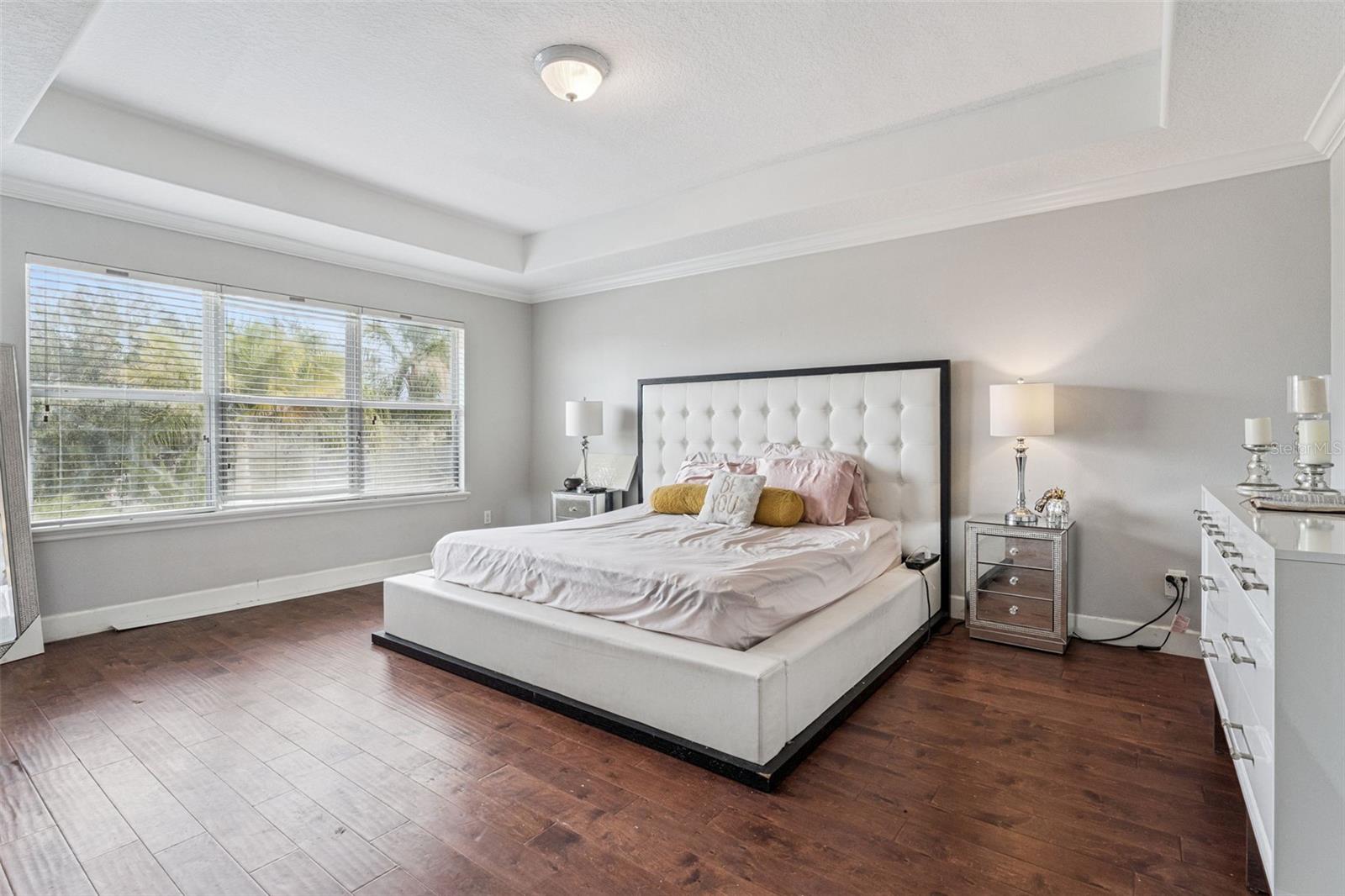Furnished bedroom with a white tufted bed, mirrored nightstands, and wide windows with blinds.