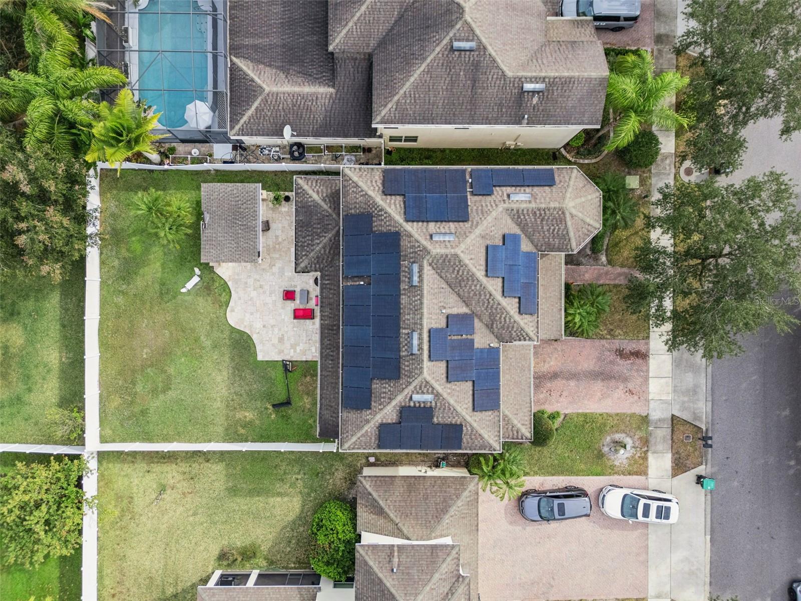 Aerial view of a home with extensive rooftop solar panels, a fenced lawn, and a stone patio.