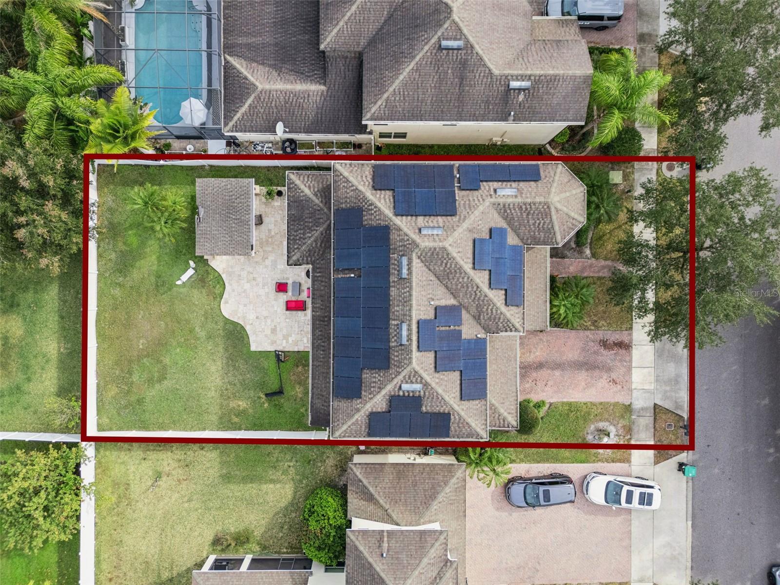 Aerial view of a home with extensive rooftop solar panels, a fenced lawn, and a paved patio.