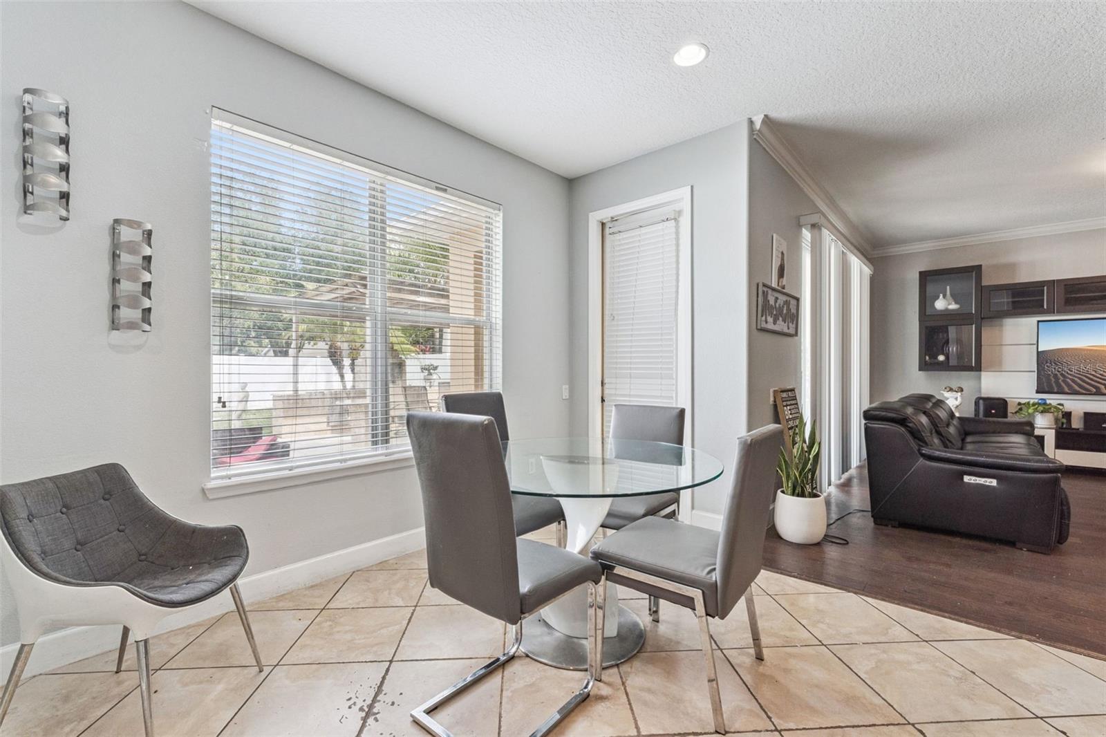 Dining area with a glass table and gray chairs beside a large window, opening to a living room.