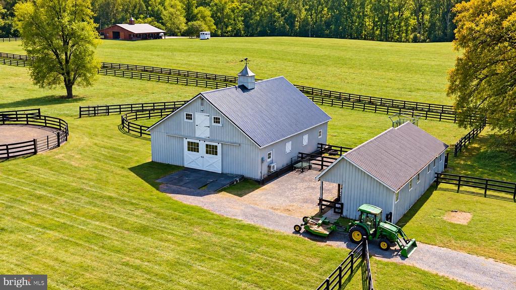 Aerial view of two gray barns and fenced green pasture with a riding arena and tractor.