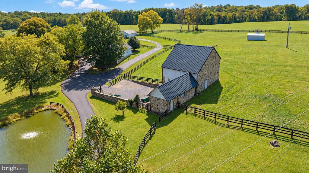 Aerial view of a stone barn beside fenced pasture, a pond, and a curved driveway.