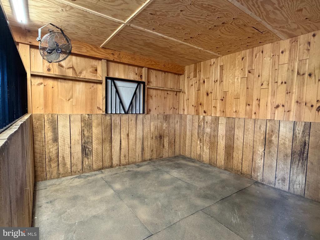 Vacant wooden horse stall with plank walls, a wall-mounted fan, and a rubberized floor.