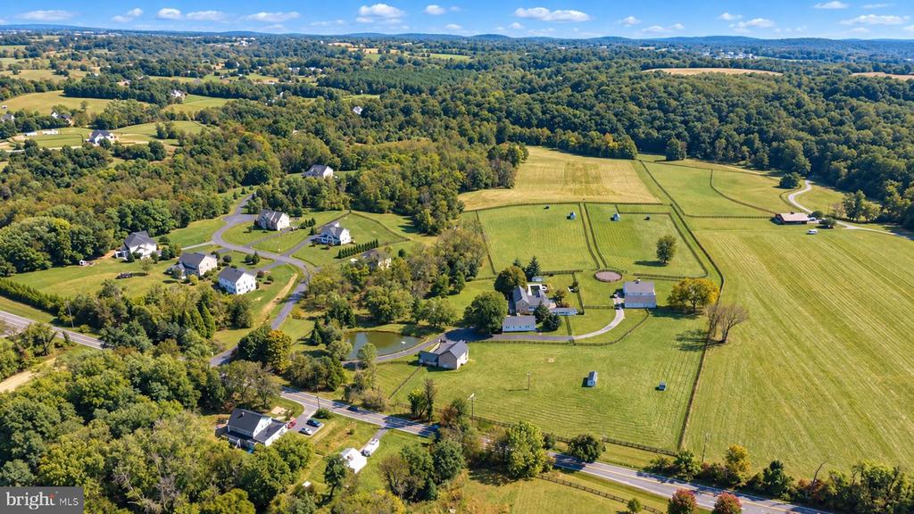 Aerial view of rural property with fenced fields, homes, a winding driveway, and wooded hills.