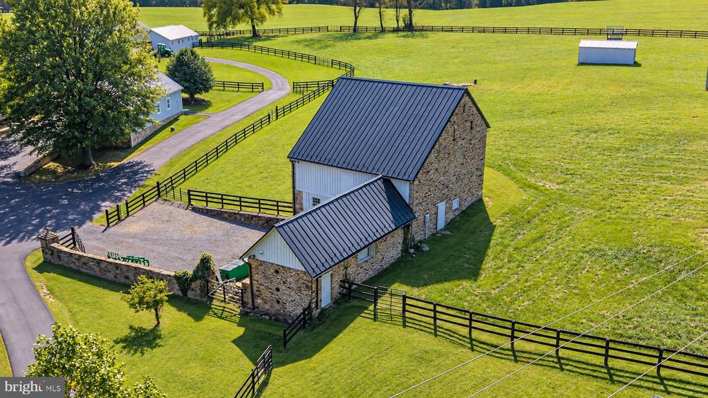 Aerial view of a stone barn with a dark metal roof, fenced pastures, and a curving driveway.