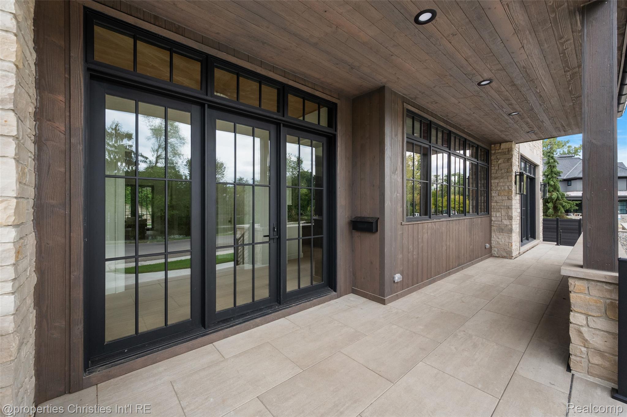 Covered porch with black-framed glass doors, wood ceiling, stone accents, and pale tiled flooring.