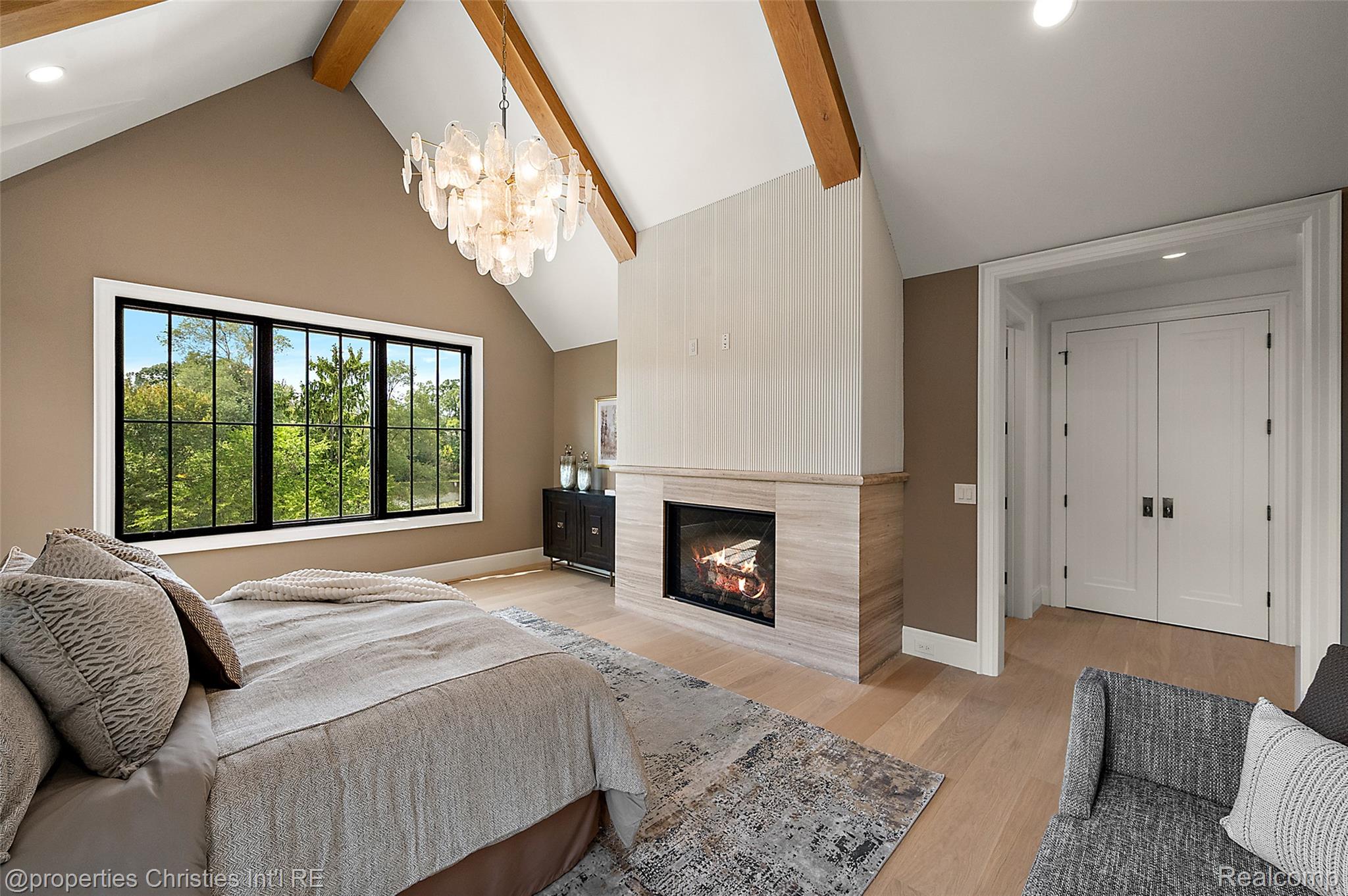 Staged bedroom with a vaulted ceiling, exposed beams, large window, and stone-surround fireplace.