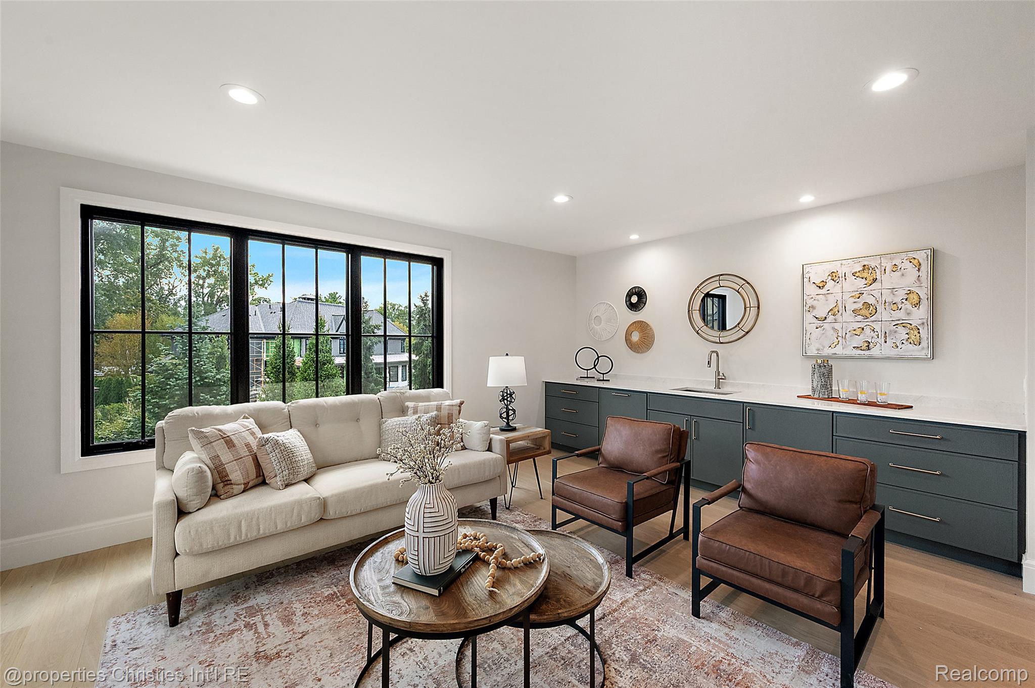 Staged living room with black-framed windows, a cream sofa, leather chairs, and a built-in cabinet with sink.