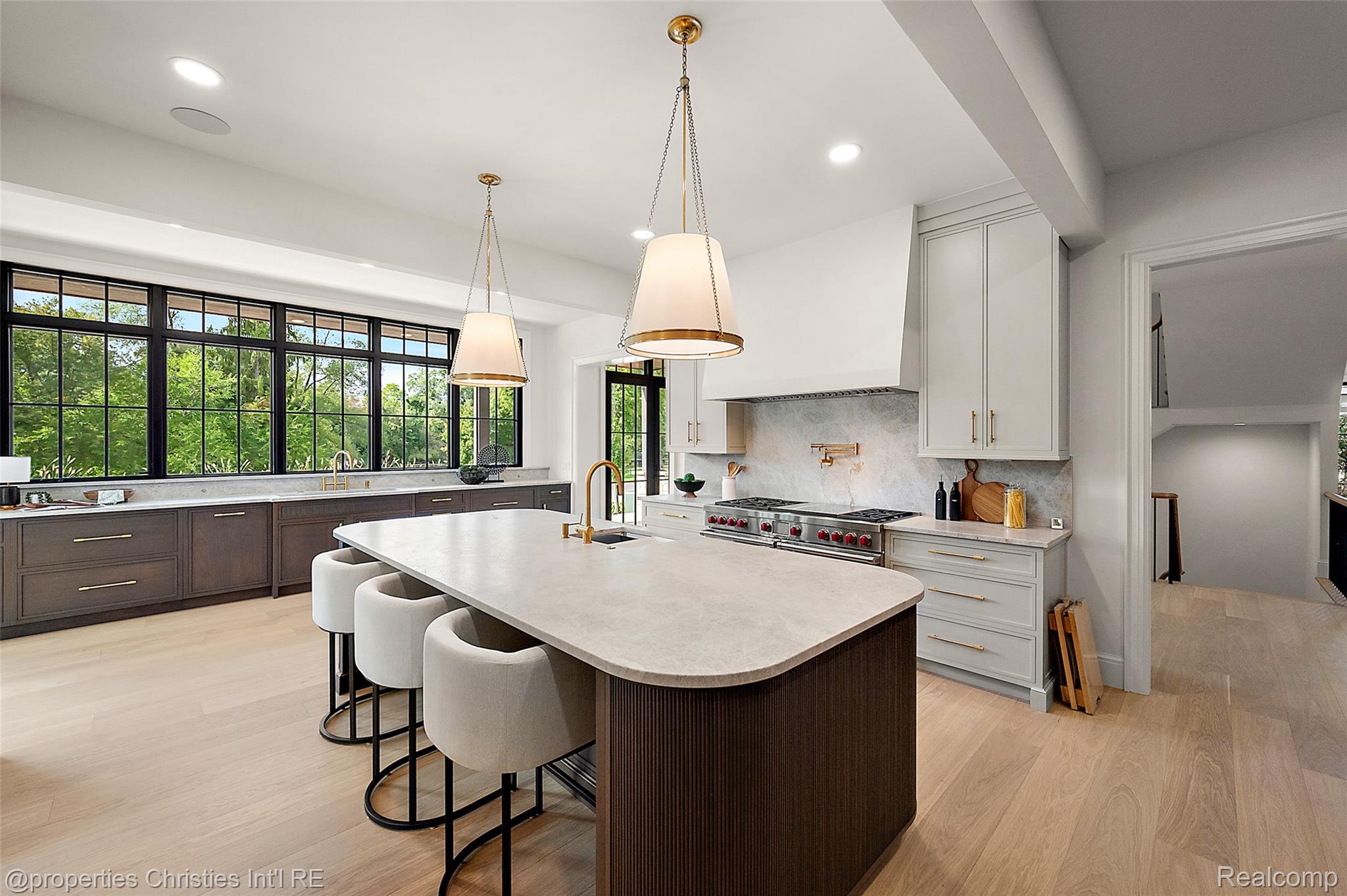 Modern kitchen with a large stone island, upholstered stools, brass fixtures, and wide windows overlooking greenery.