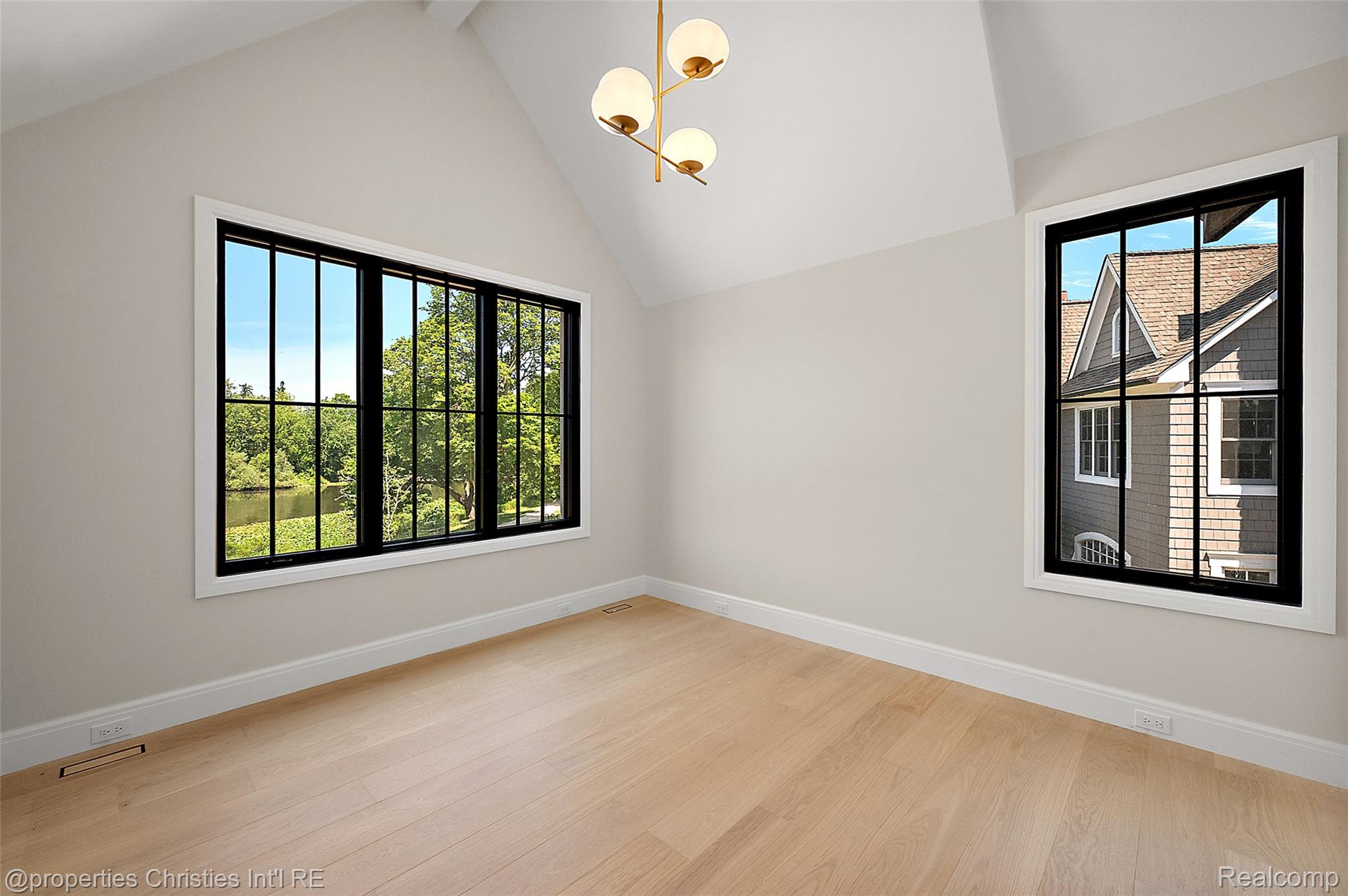 Empty room with pale wood floors, vaulted ceiling, and black-framed windows overlooking a pond and neighboring house.