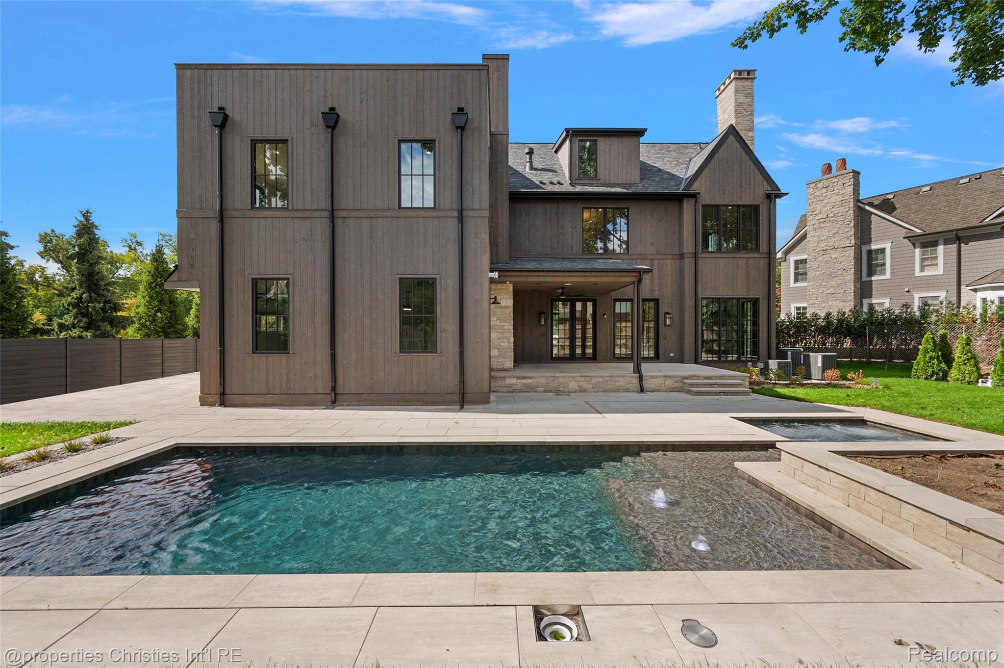 Modern dark-sided home behind a rectangular pool and broad stone patio.