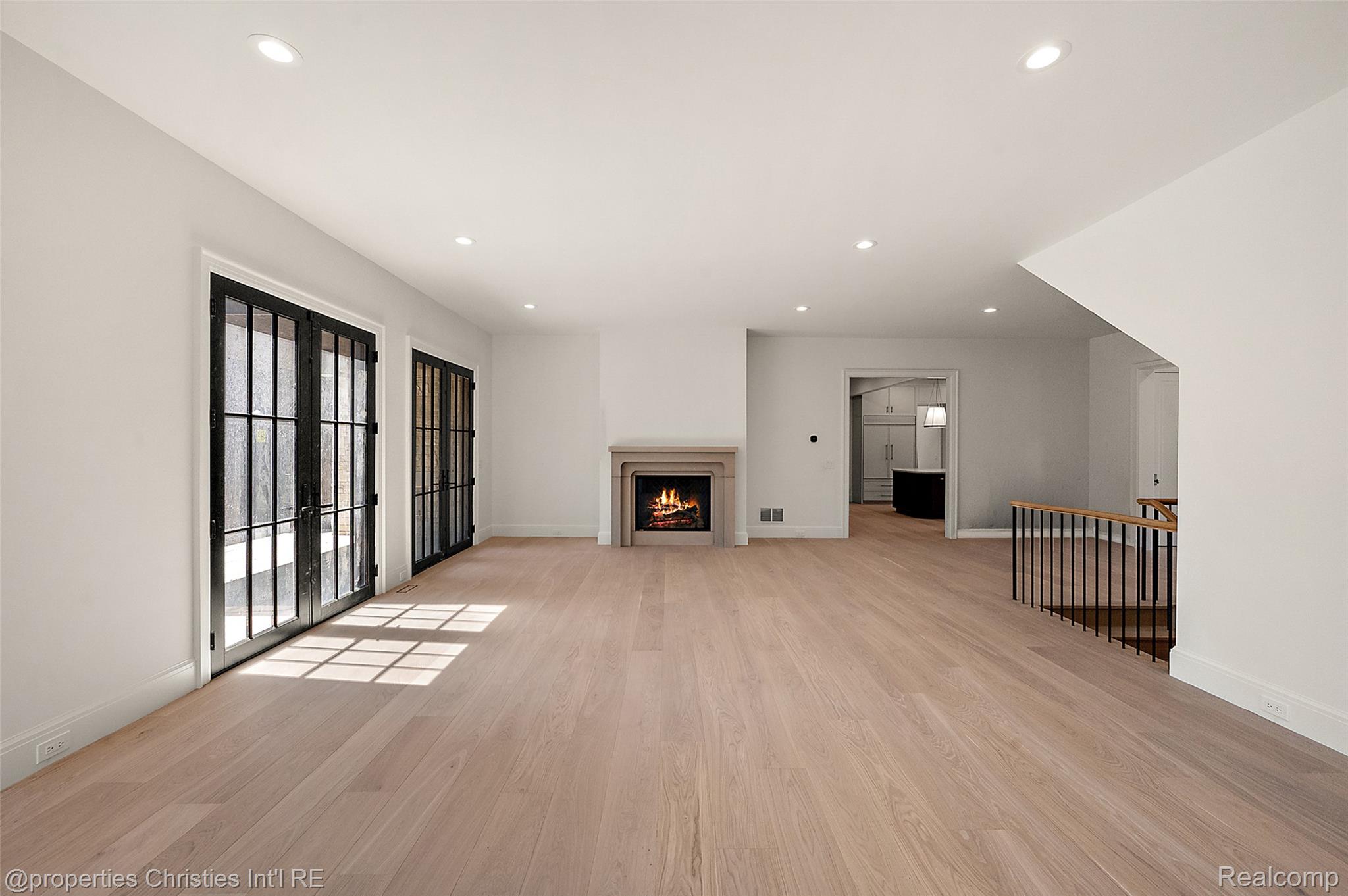 Empty living room with pale wood floors, black-framed glass doors, and a lit fireplace.