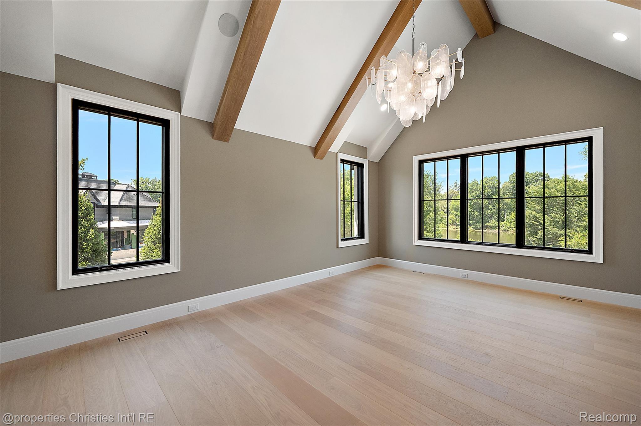 Vacant vaulted room with exposed wood beams, a chandelier, and large windows overlooking trees and water.