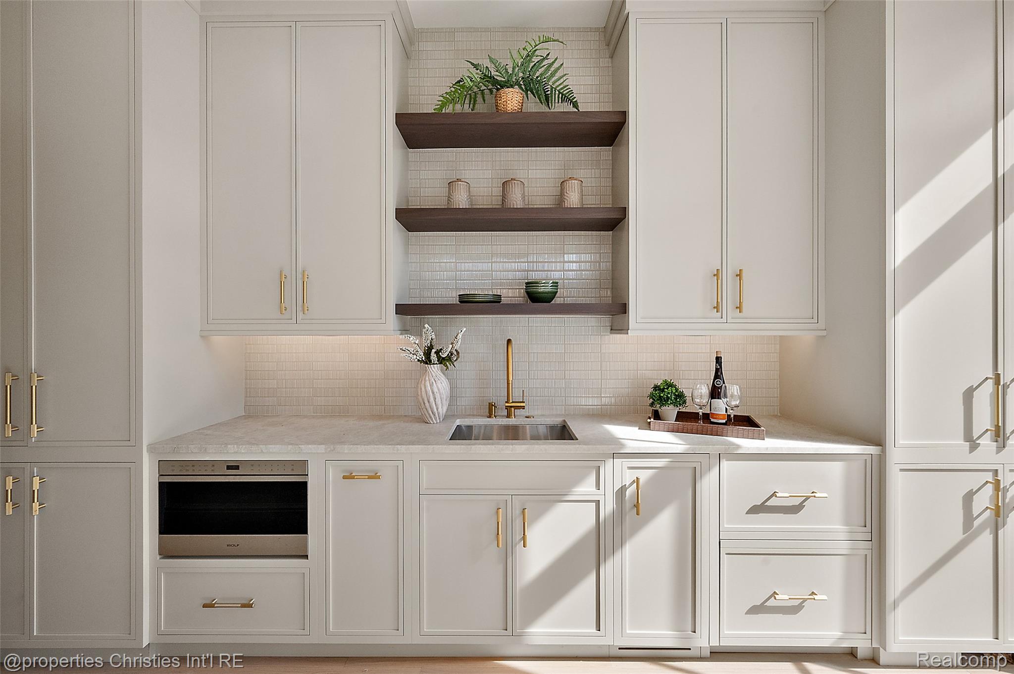 Staged kitchen with pale gray cabinets, brass hardware, floating shelves, and a tiled backsplash.