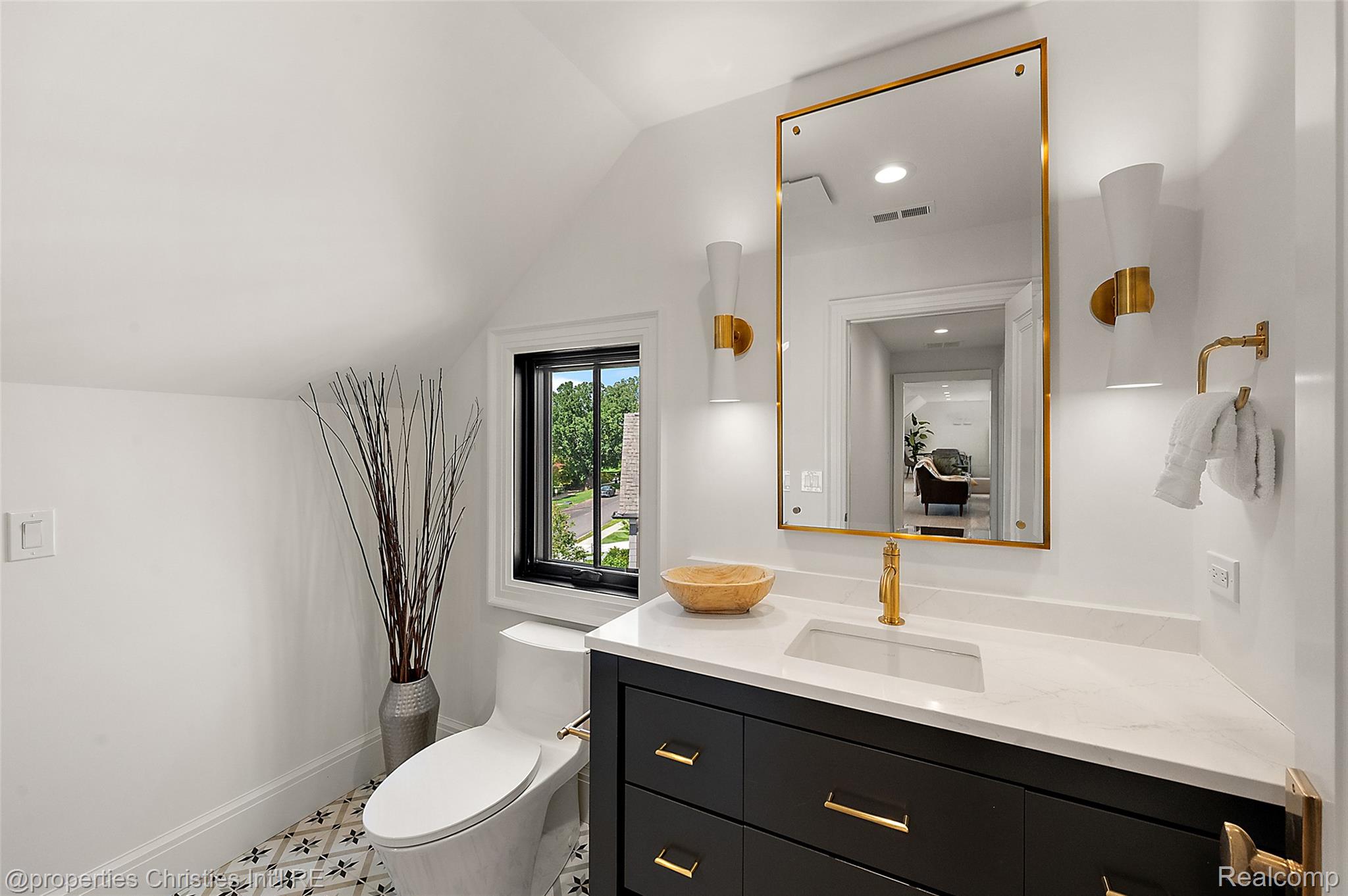 Bright bathroom with a black vanity, gold fixtures, patterned tile floor, and a window overlooking greenery.
