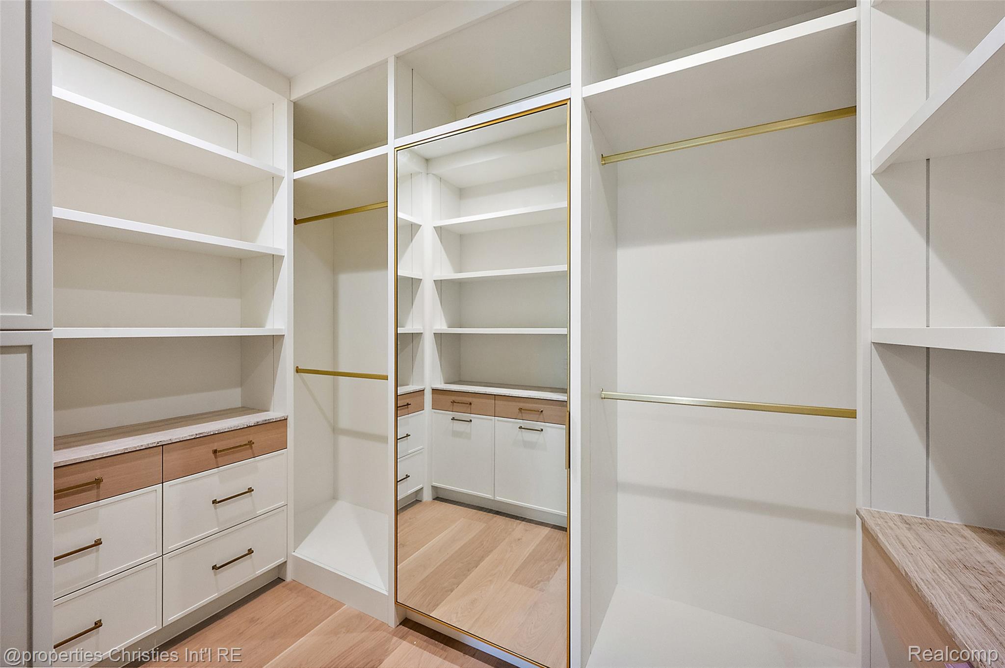 Walk-in closet with built-in white shelving, brass rails, wood-front drawers, and a full-length mirror.