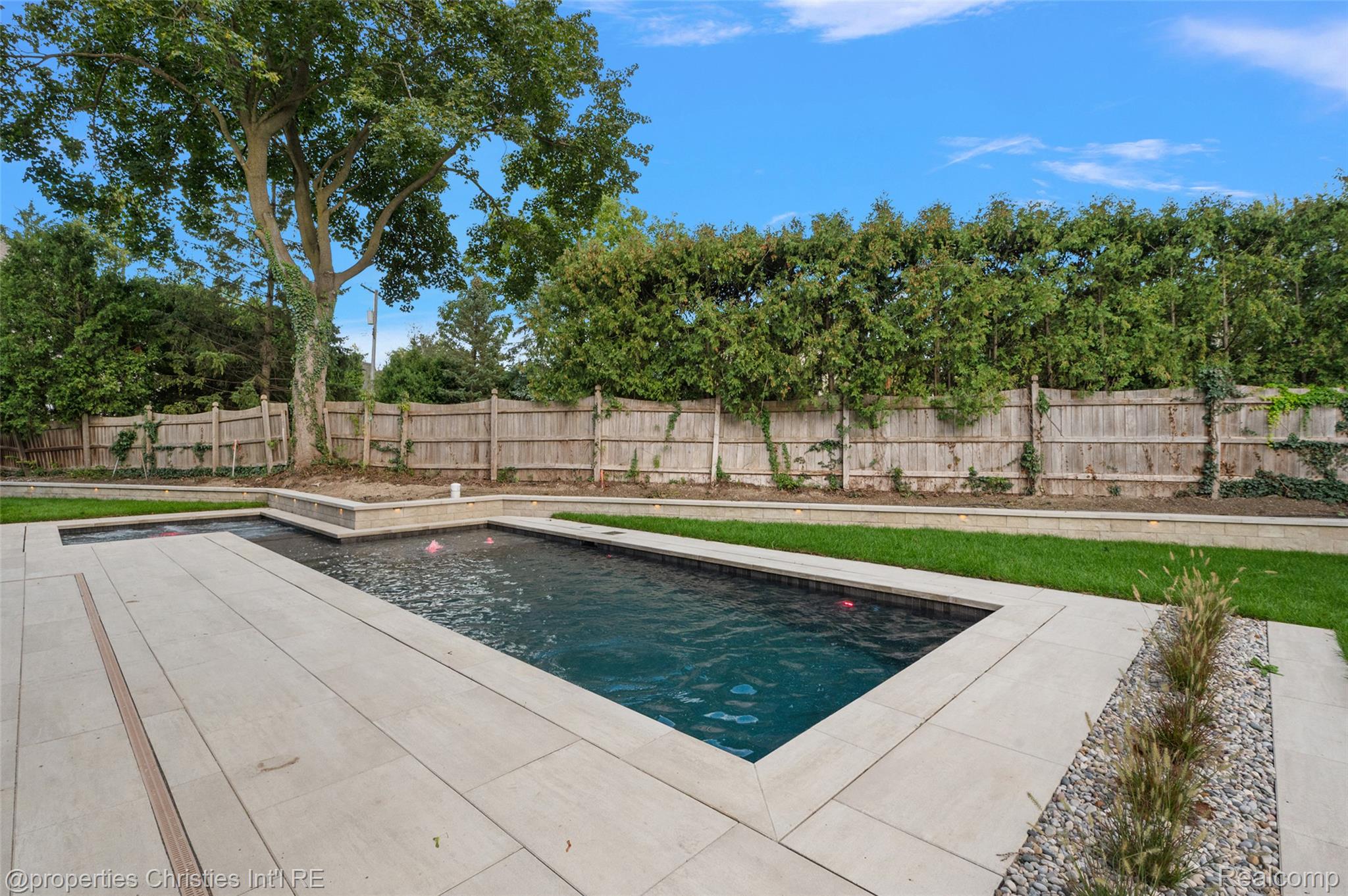 Backyard swimming pool with pale stone paving, a wooden privacy fence, and mature greenery.