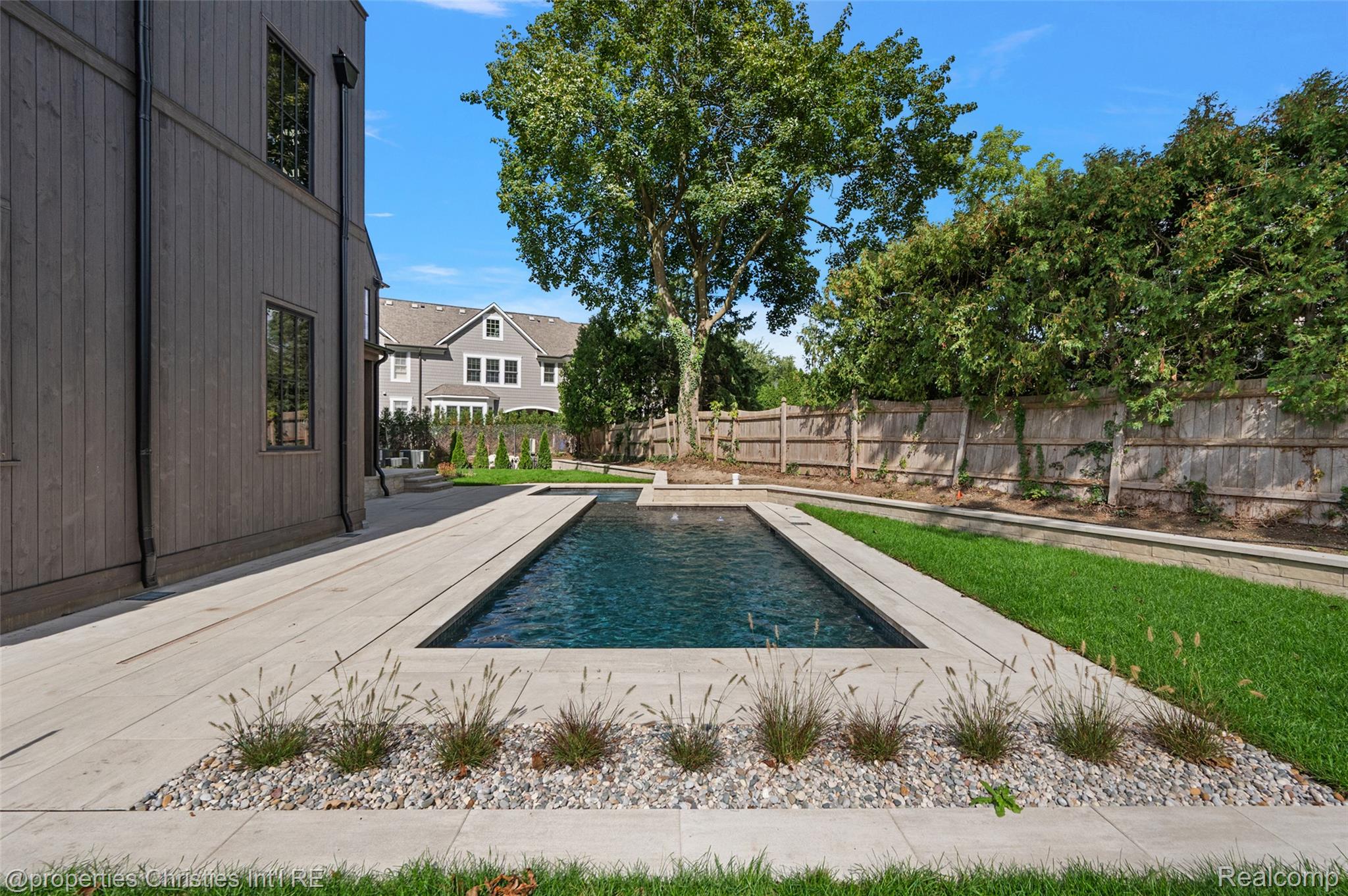 Rectangular backyard pool beside a dark-sided house, bordered by a pale stone deck and landscaped lawn.