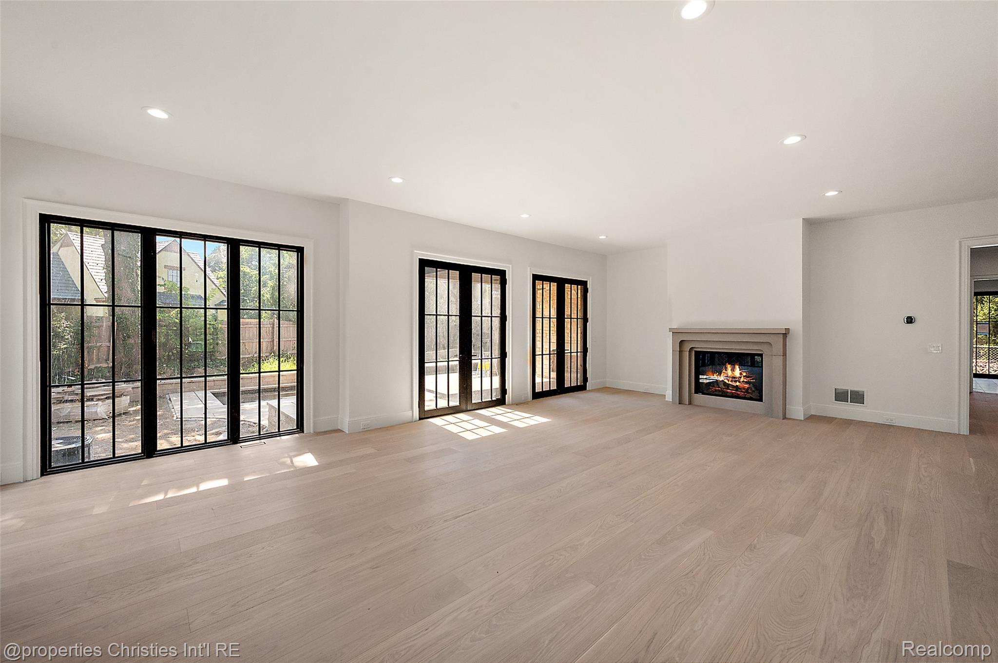 Empty living room with pale wood floors, black-framed glass doors, and a lit fireplace.