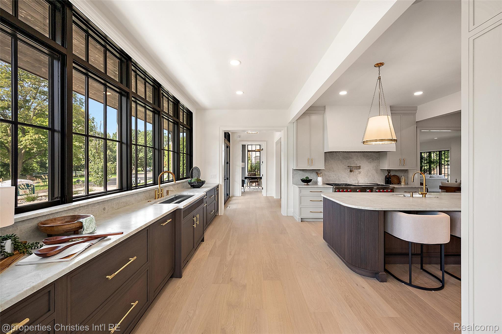 Bright kitchen with a curved island, brass fixtures, and tall black-framed windows overlooking trees.