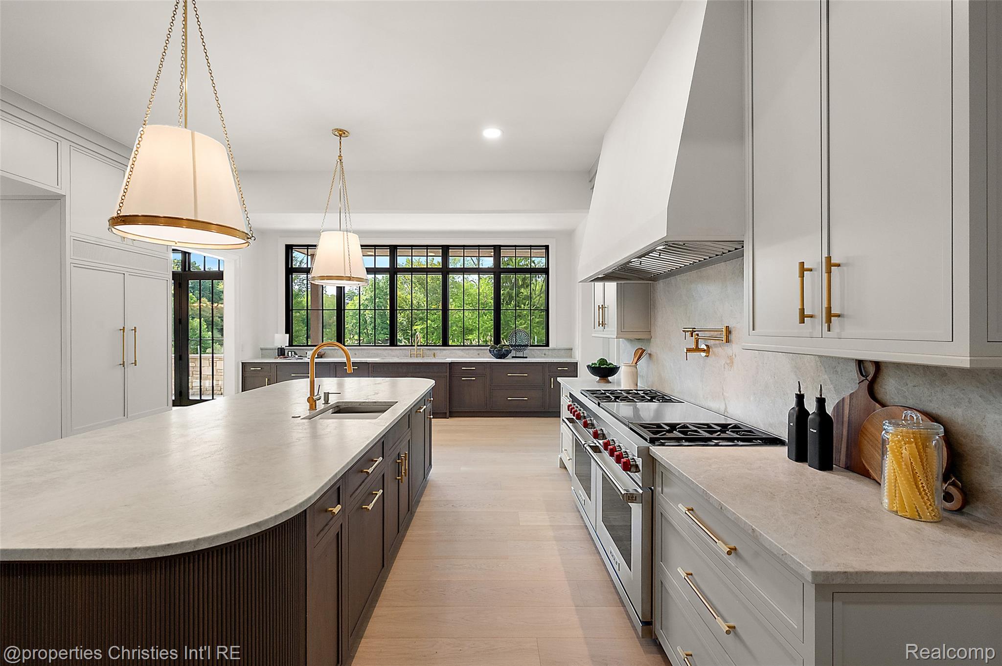 Modern kitchen with a large island, brass fixtures, professional range, and black-grid windows overlooking greenery.