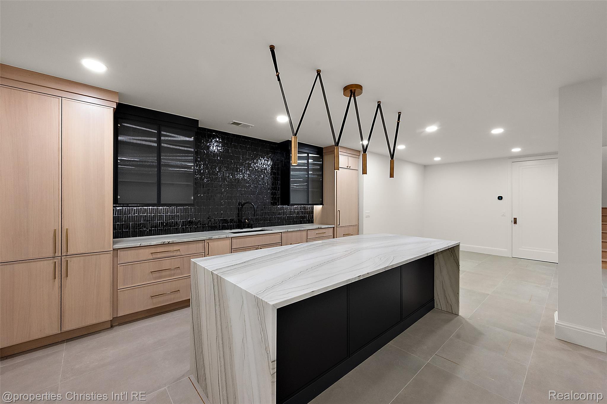 Modern kitchen with a veined stone island, pale wood cabinets, black tile backsplash, and geometric pendant lights.