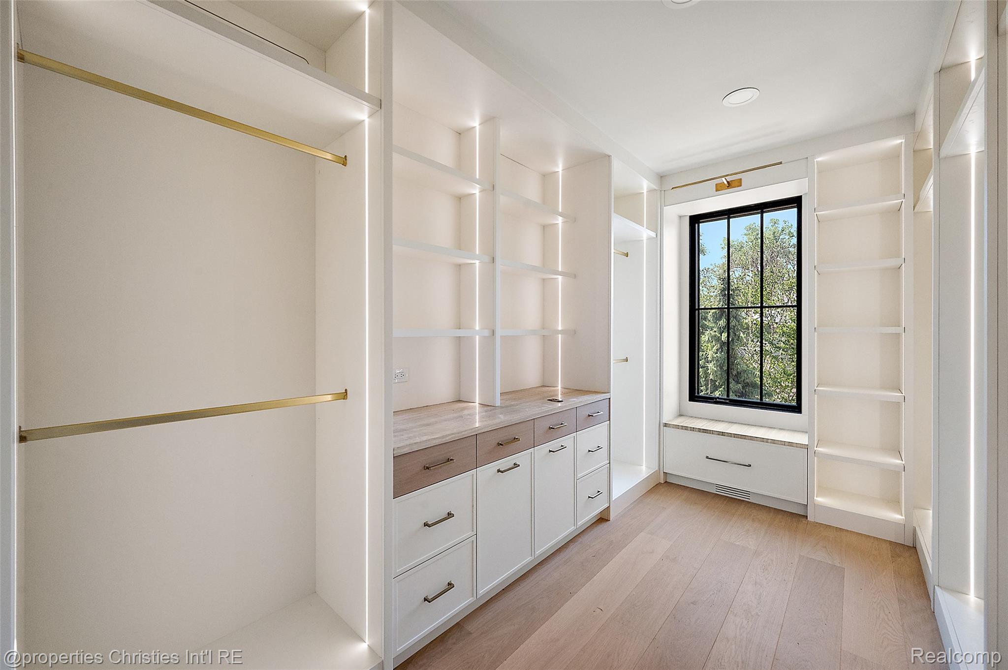 Walk-in closet with built-in shelving, brass rods, wood-front drawers, and a window seat.